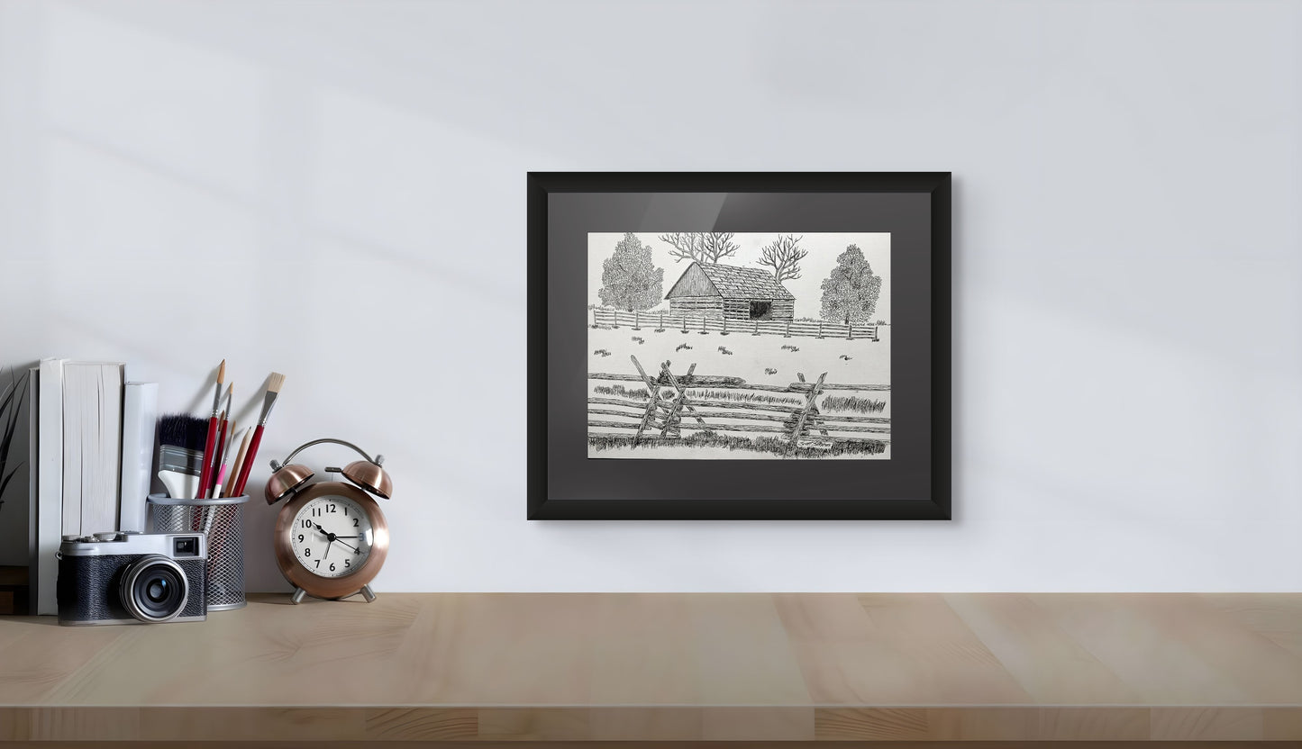 Framed black and white print of a landscape with buildings on a desk against a white wall.