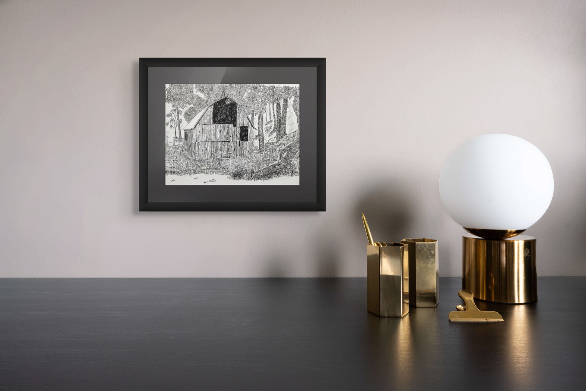 Framed black and white drawing of old barn  on a wall above a desk with decorative items.