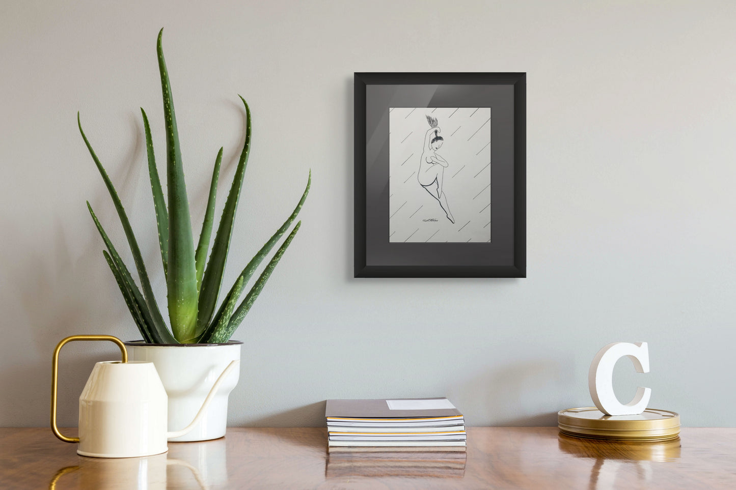 Framed artwork on a wall above a desk with a plant and decorative items.