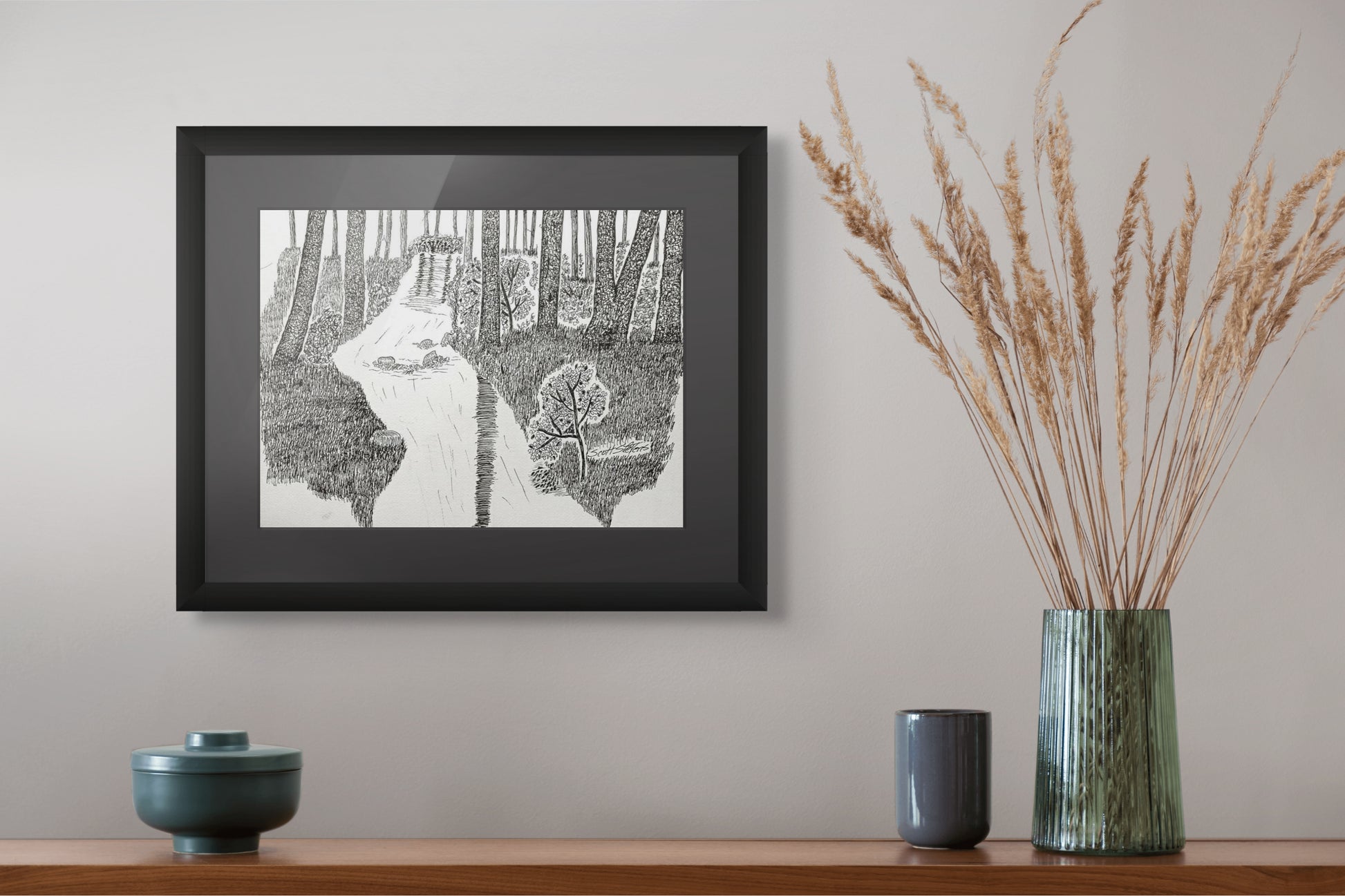 Framed black and white artwork of a forest scene on a wall above a wooden shelf with decorative items.