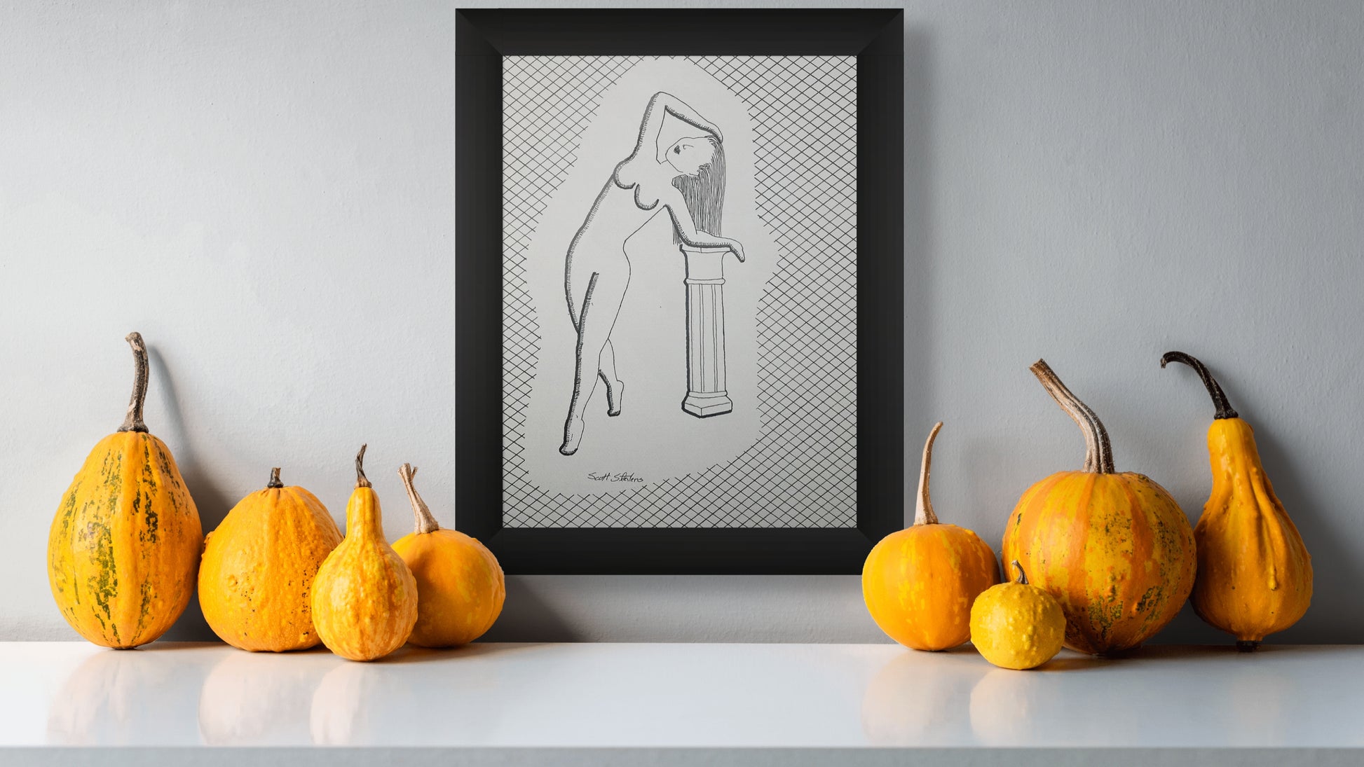 Framed artwork of a nude figure on a gray wall with pumpkins and gourds on a white surface.