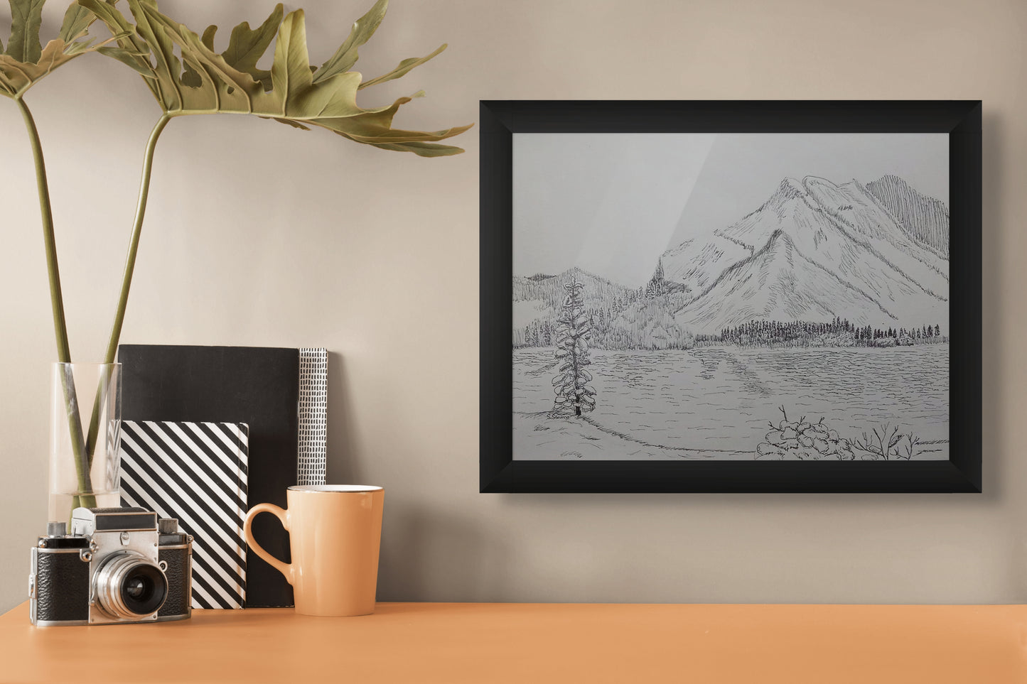 Framed black and white mountain landscape drawing on a wall above a desk with a camera, mug, and books.