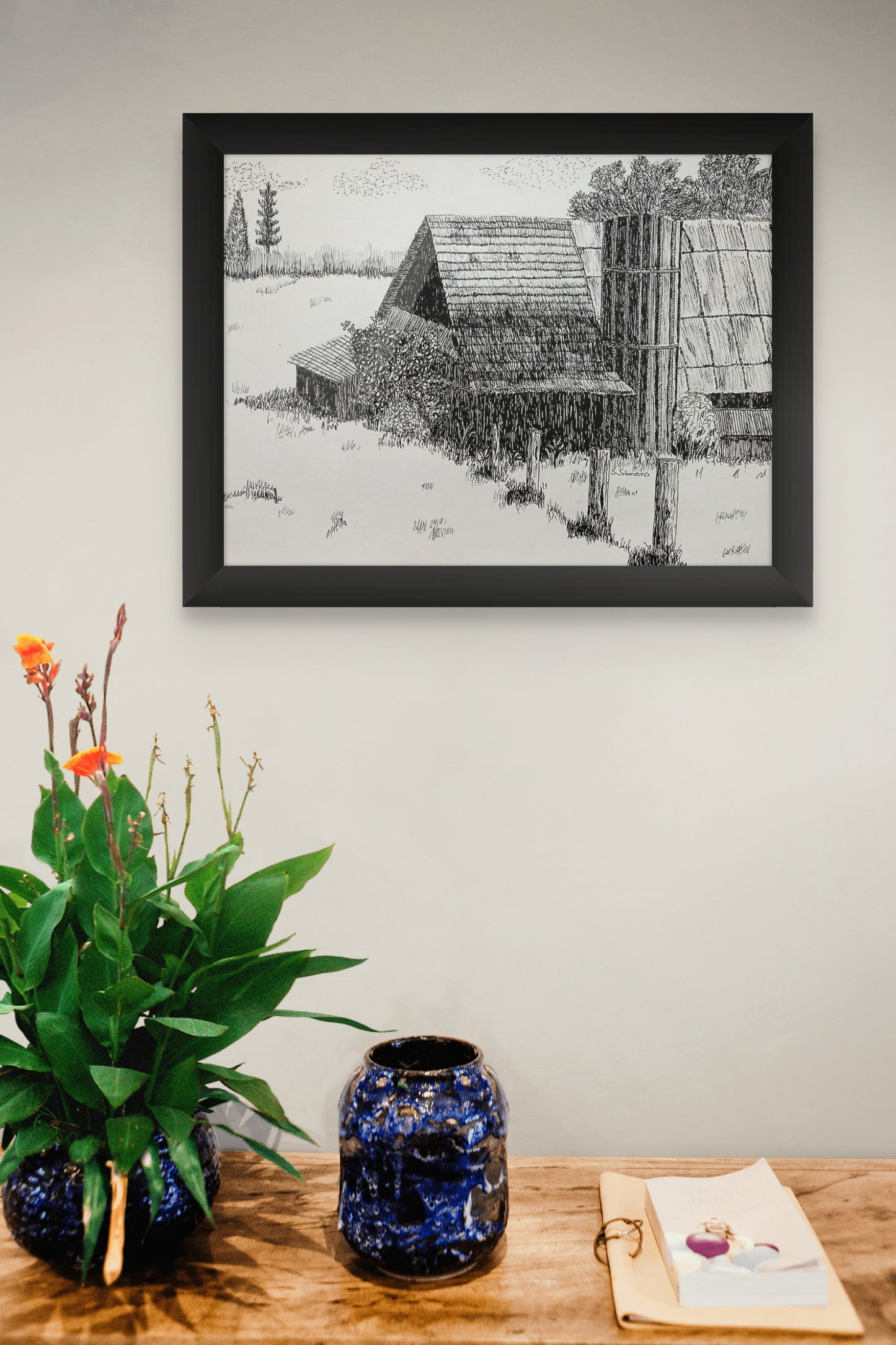 Framed black and white artwork of a landscape with a cabin on a wall above a wooden table with decorative items.