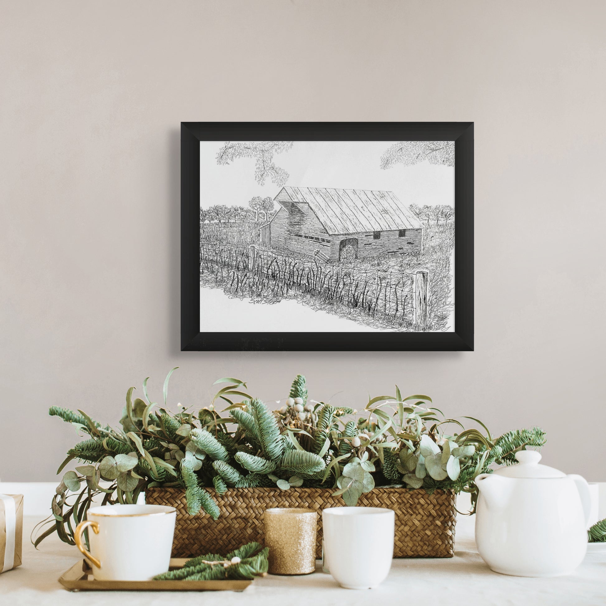 Framed black and white artwork of a barn above a decorative table setting with plants and teacups.