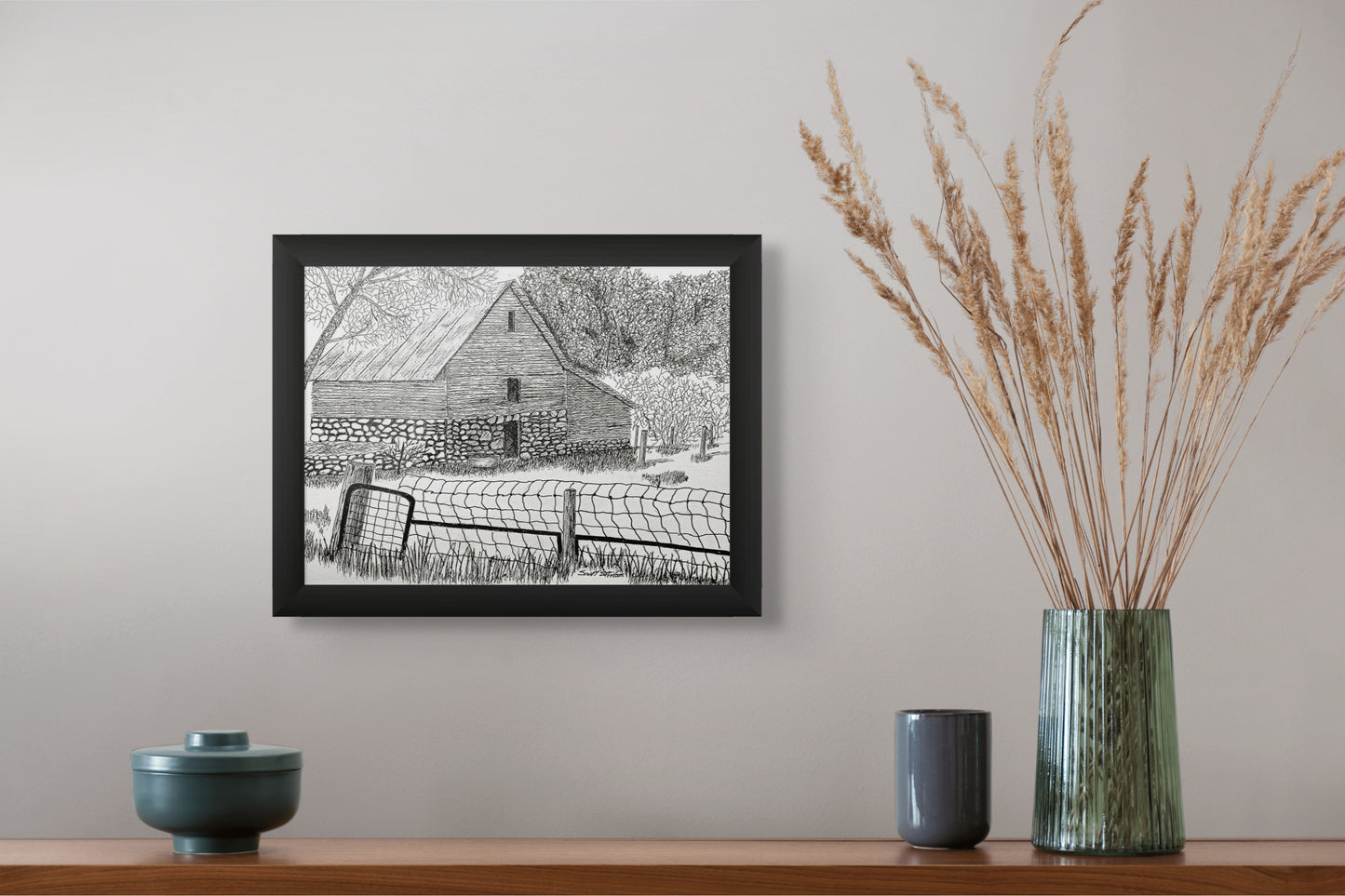 Framed black and white print of a barn on a wall above a wooden shelf with decorative items.