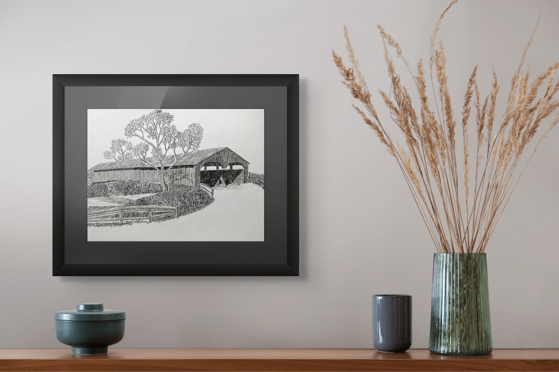 Framed black and white landscape print on a wall above a wooden shelf with decorative items.