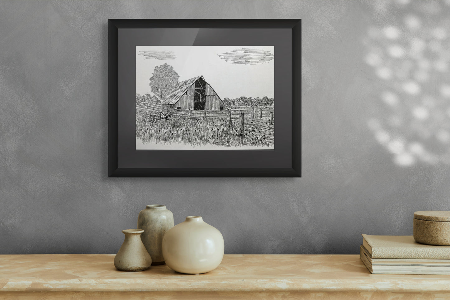 Framed black and white print of a barn on a gray wall above a wooden surface with decorative items.