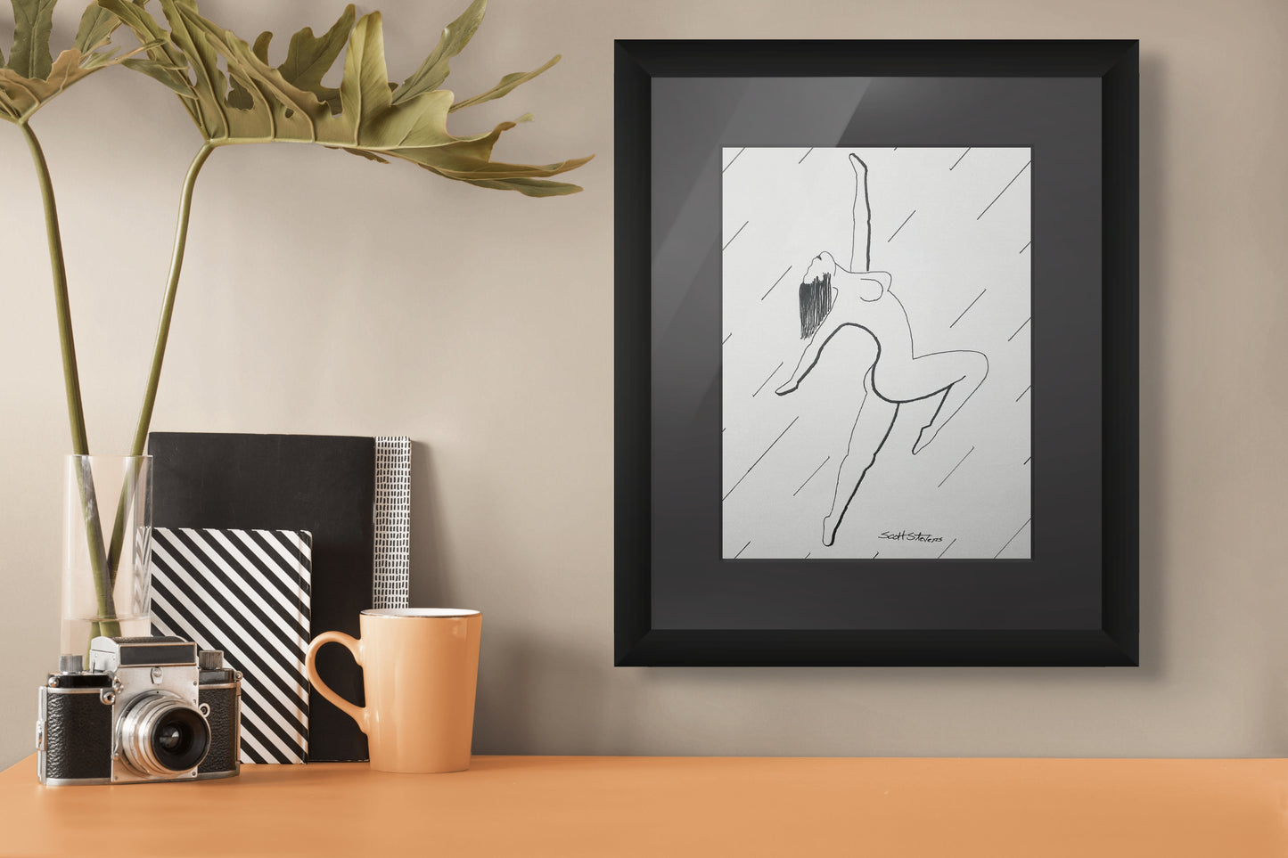 Framed pen and ink artwork of a nude woman dancing on a wall with a camera, mug, and books below.