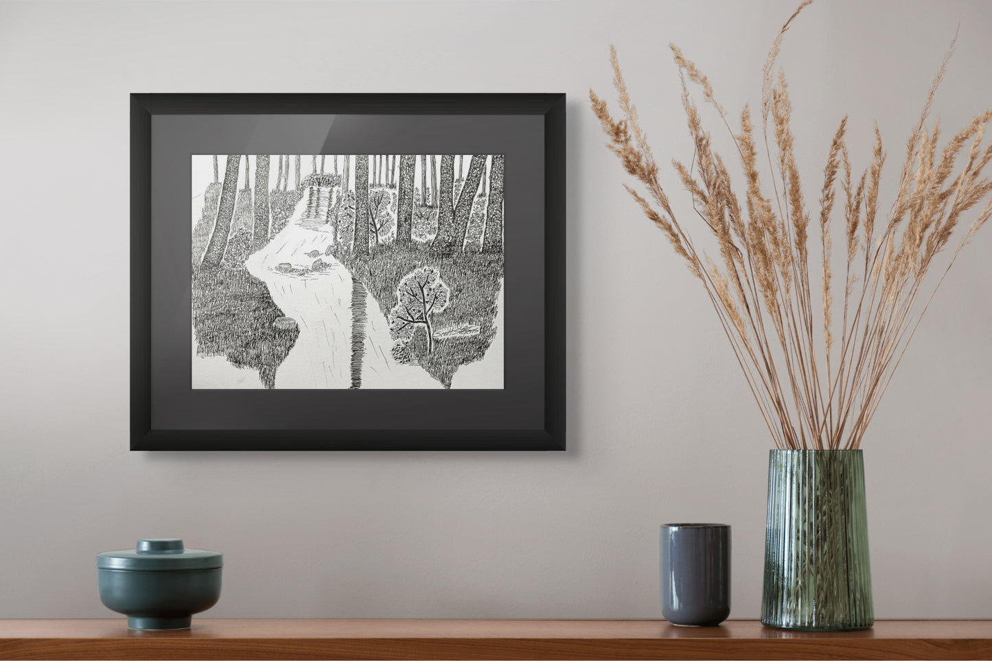 Framed black and white artwork of a forest scene on a wall above a wooden shelf with decorative items.