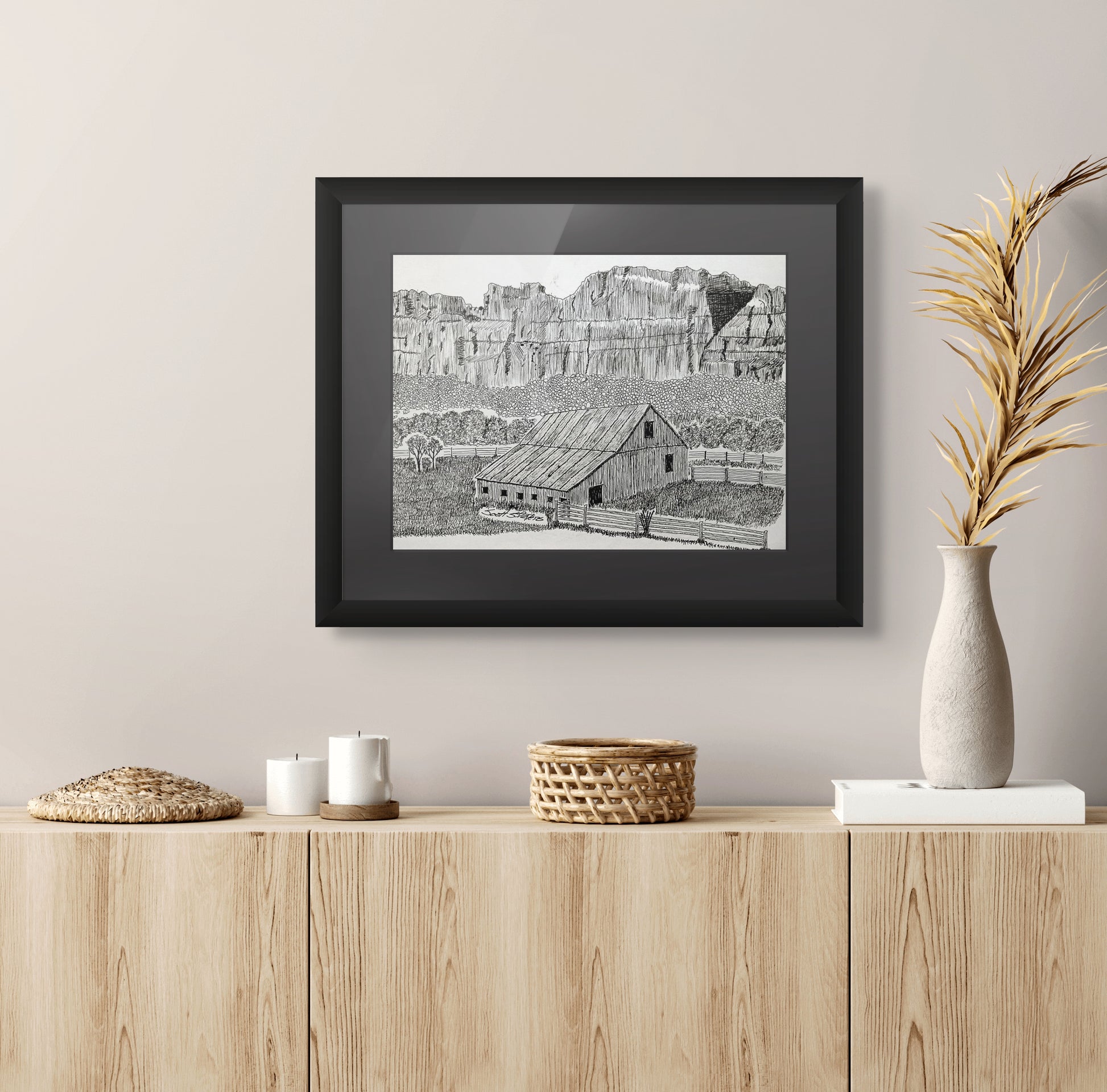 Framed black and white print of a landscape on a wall above a wooden cabinet with decorative items.