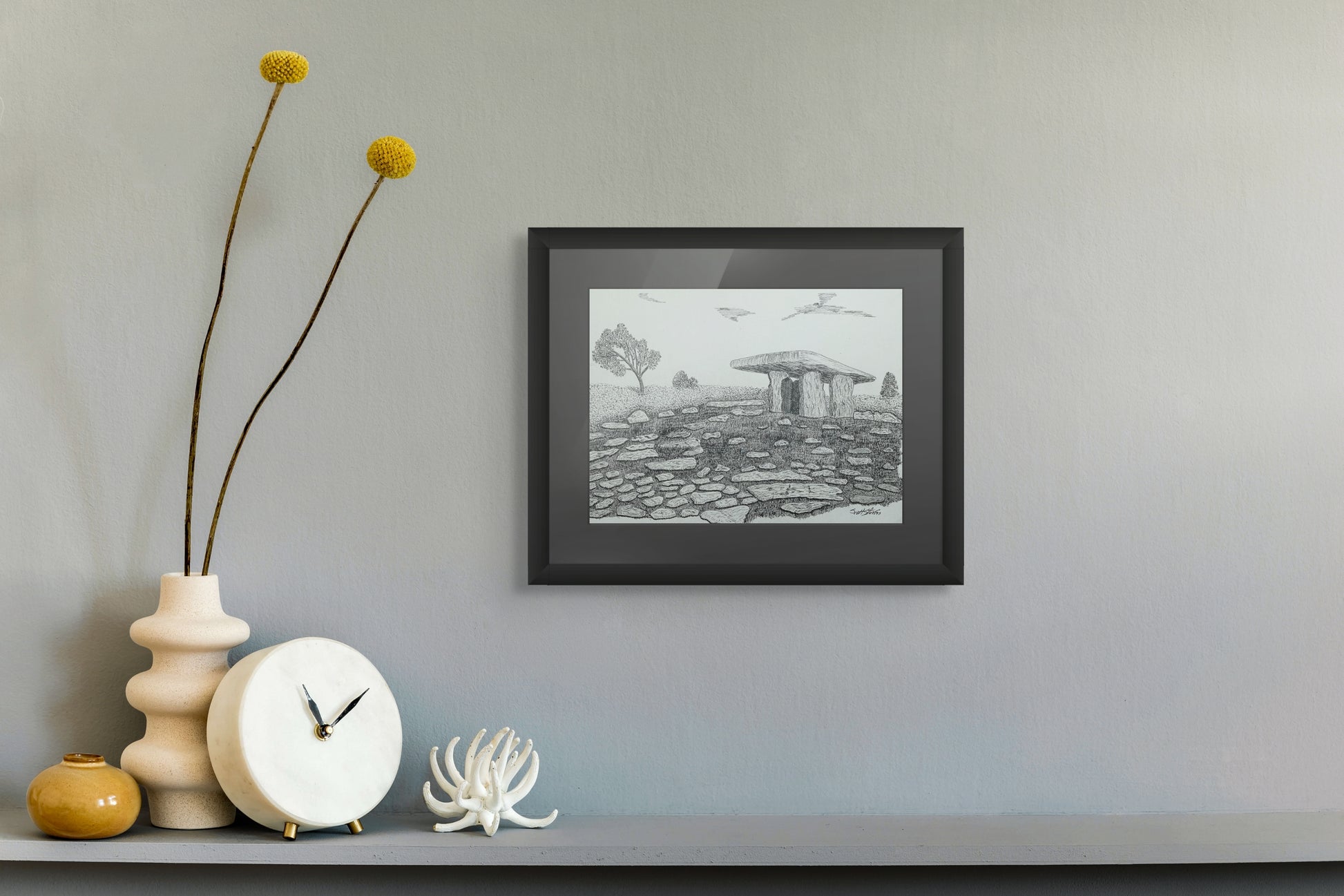 Framed black and white print of a landscape on a wall with decorative items below.